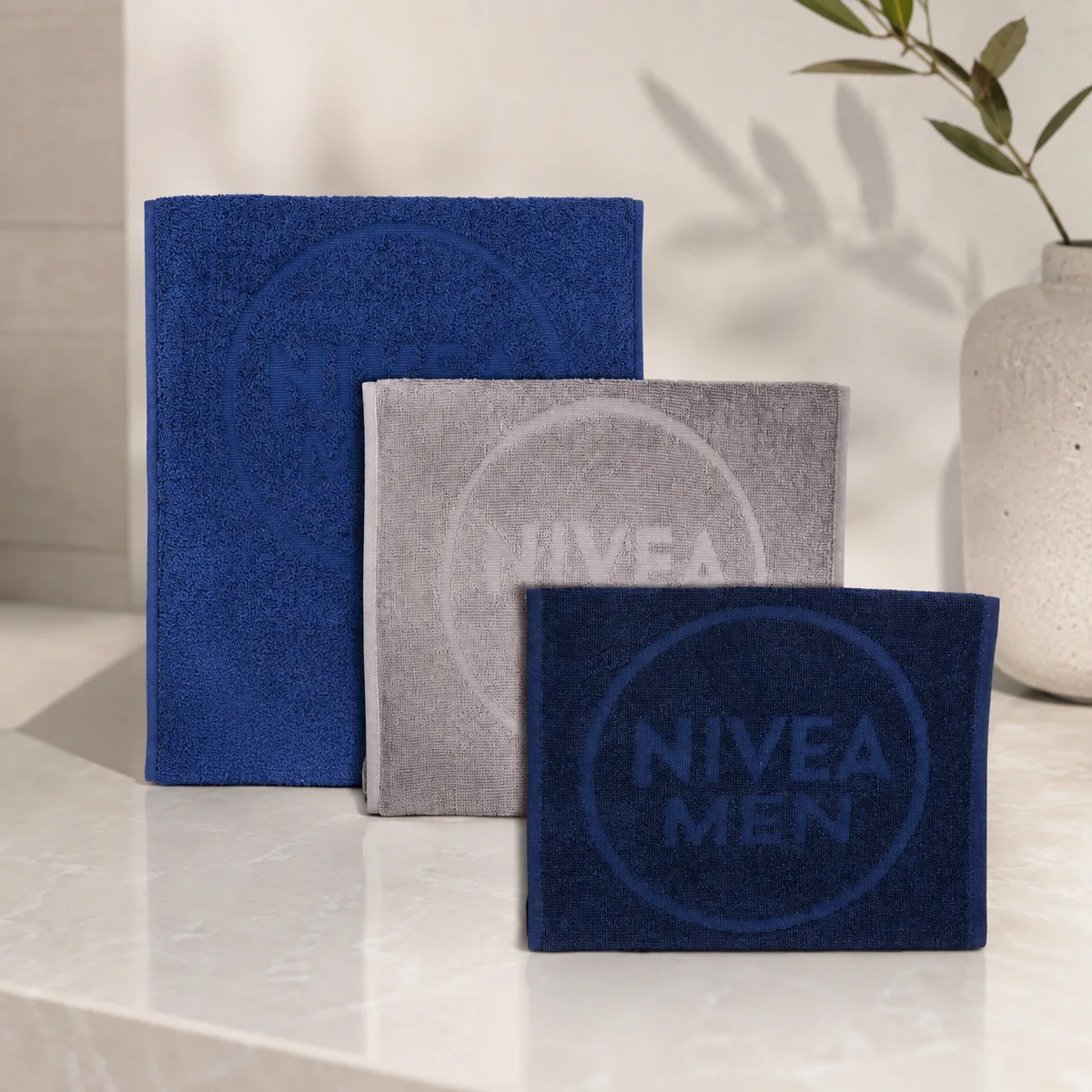 Három összehajtott frottír törölköző – kék, szürke és sötétkék – Nivea Men emblémával, világos pulton